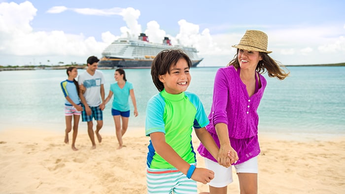 Uma família de cinco pessoas passeia em uma praia com um navio da Disney Cruise Line atracado ao fundo.
