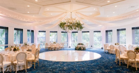 An elegant reception setup with round tables, floral centerpieces and draped fabric under a sparkling chandelier