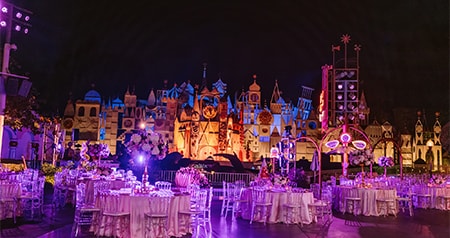 An evening reception setup with round tables and chairs in front of the illuminated 'it’s a small world' facade