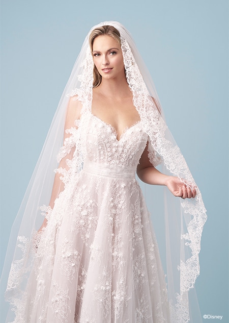 A bride wearing a lace edged veil and a Snow White inspired A line wedding gown featuring a V neckline, embroidery, spaghetti straps and lace off the shoulder sleeves