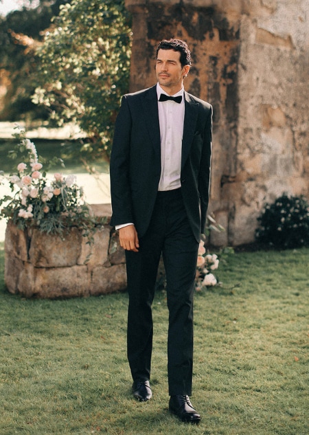 A man in a black tuxedo and bow tie walking across a lawn near a stone archway