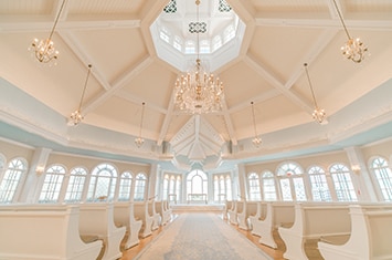 The Disney Wedding Pavilion at Walt Disney World Resort with 2 rows of pews with an aisle leading towards an altar beneath a round ceiling with hanging lights