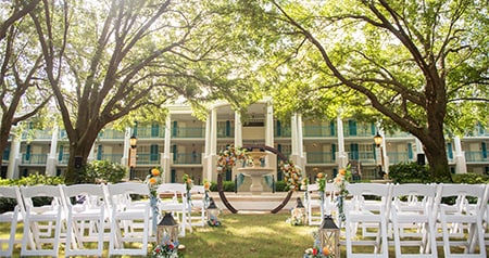 An outdoor wedding ceremony setup in front of a hotel with classic wedding chairs, floral accents and a circular arch under large trees