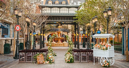 An outdoor ceremony setup with floral arches, wooden chairs, a decorative cart and a fountain