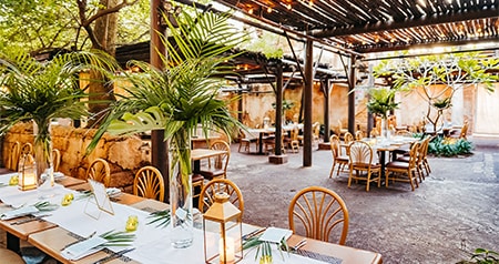 An outdoor dining setup with long tables, tropical centerpieces, lanterns and a rustic pergola with string lights