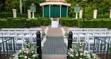 An outdoor wedding ceremony venue with a gazebo, altar, floral arrangement, hedges and rows of chairs at the United Kingdom Courtyard at Epcot