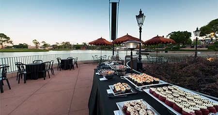 An outdoor wedding reception space with a garden, buffet tables, lampposts and tables with chairs next to a lake at United Kingdom Lochside at Epcot