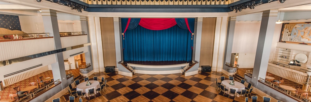 A ballroom with a checkered dance floor in front of a stage surrounded by many tables and chairs