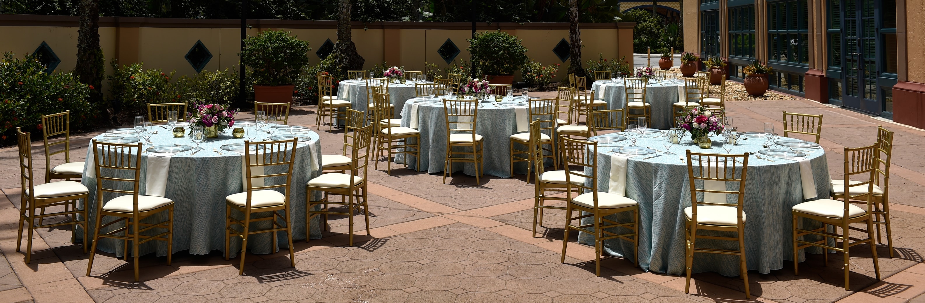 Five circular tables with chairs arranged in a courtyard surrounded by plants