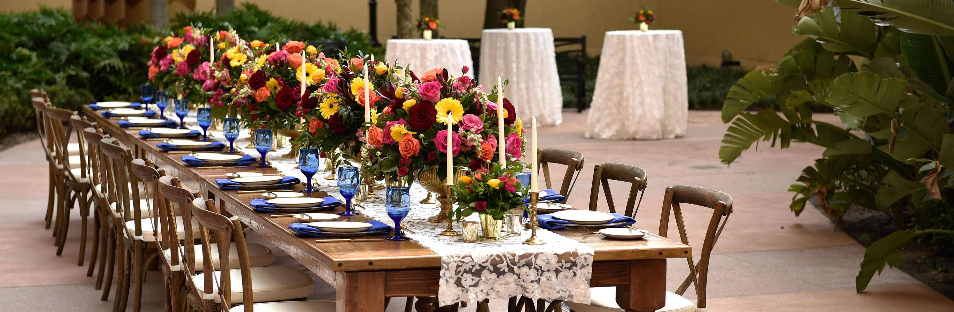 A long table with many place settings on a courtyard with 3 table tops in the background