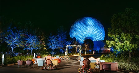 An outdoor wedding ceremony venue with rows of upholstered furniture and a floral arch, surrounded by gardens with Spaceship Earth in the background