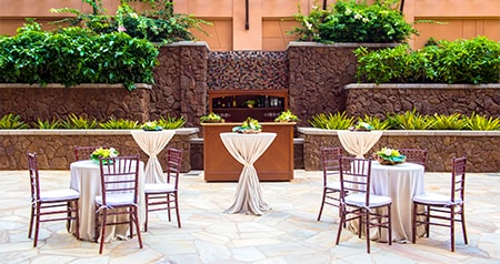 Dining and cocktail tables decorated for a wedding in Kipuka Courtyard, against a rustic, stone wall backdrop