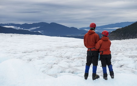 A couple suited up in mountaineering gear stand on an ice field enjoying the Alaskan expanse
