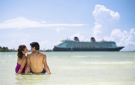 A man and a woman in bathing suits sit on the beach with a Disney Cruise Line ship in the background
