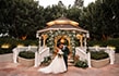 A bride and groom embracing and laughing while standing in front of a gazebo featuring hedges, a rose trellis and a chandelier