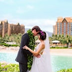 A newlywed bride and groom wearing a flower crown and lei while embracing on a beach with Aulani, A Disney Resort and Spa in the background