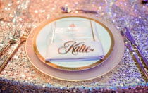 A single plate setting on a sequined tablecloth with a wedding invitation and the name, ‘Katie’ 