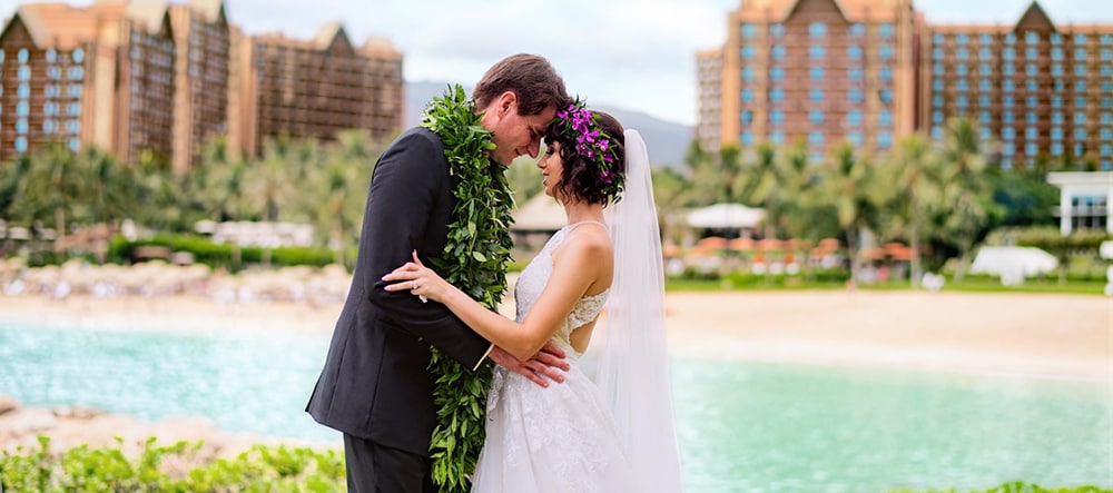 A man and a woman, draped in a Hawaiian lei and headdress, hold each other and stand nose to nose with Aulani Resort in the background