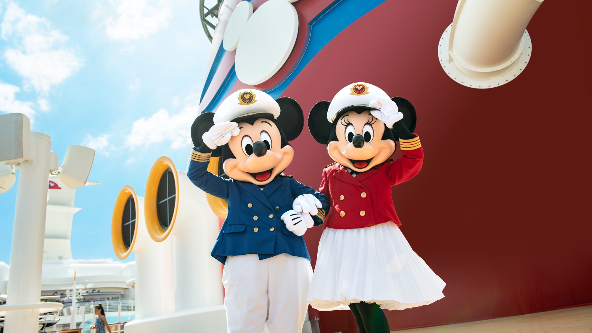 Alt テキスト：ディズニー・クルーズラインの客船のデッキに立ち、腕を取り合って挨拶するミッキー船長とミニー船長