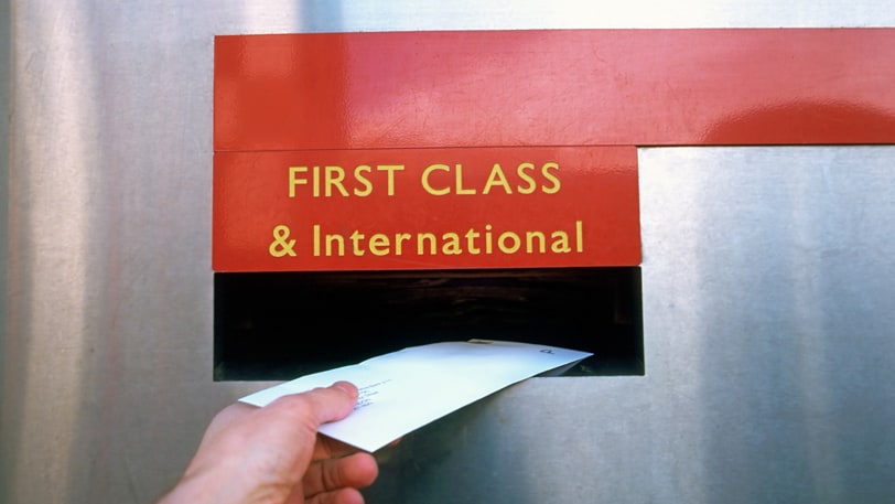 A man's hand puts a letter in a mailbox with an engraved sign reading First Class & International