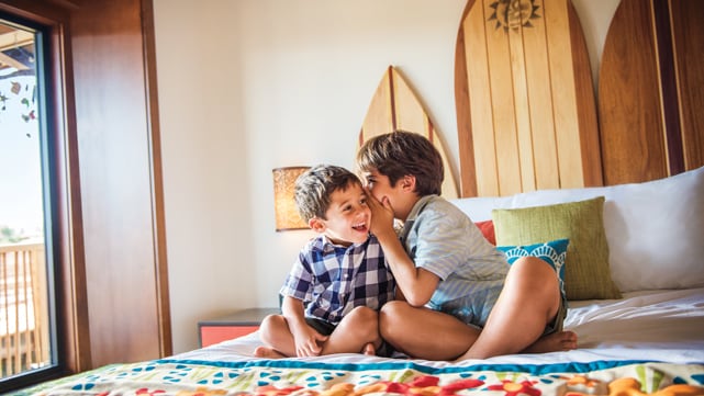 Two young brothers share a secret while sitting on a bed with a surfboard-styled headboard