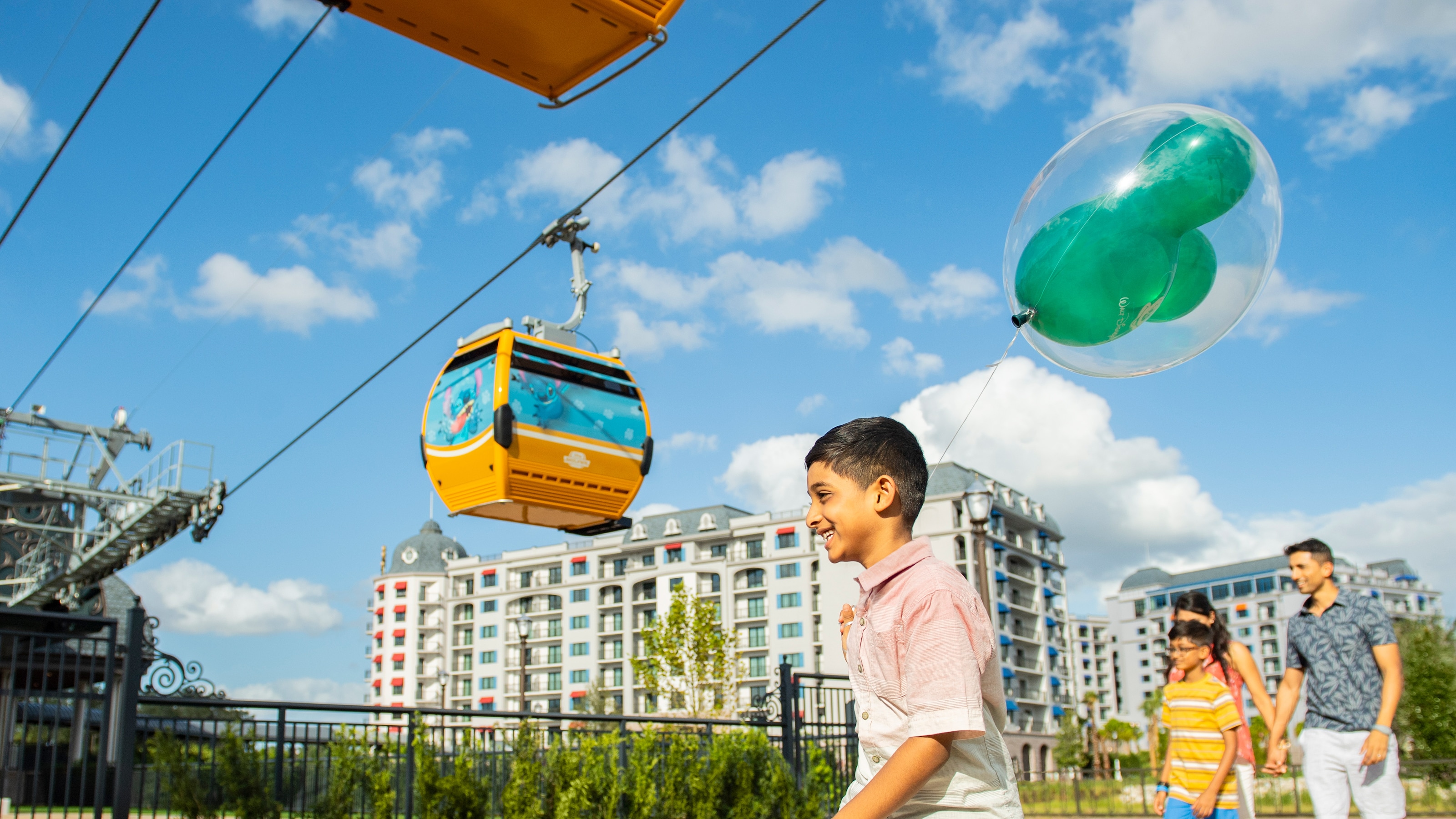 A family of 4 walking below a Disney Skyliner aerial gondola near Disney’s Riviera Resort