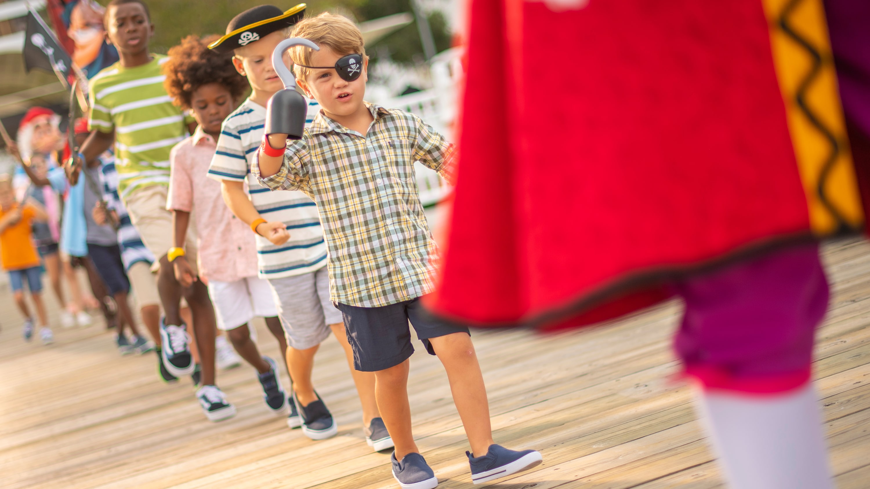 Children wearing pirate hats, hooks or eye patches follow Captain Hook on a boardwalk, parade style