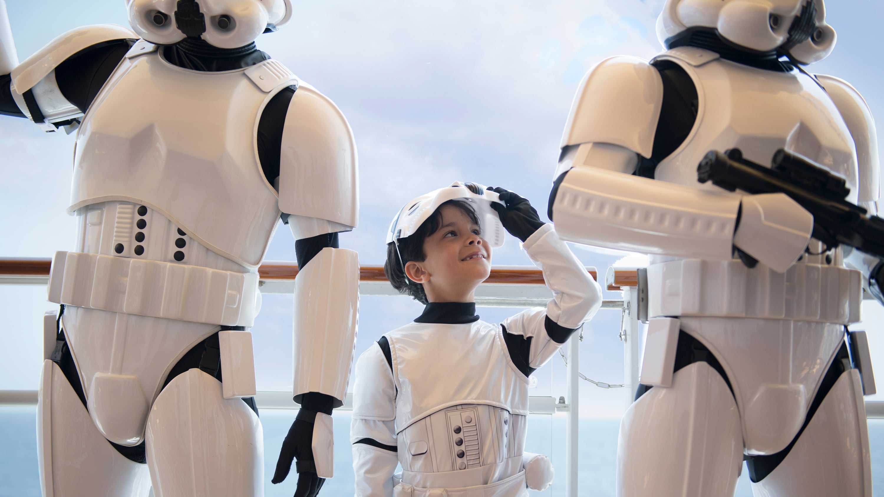 A young boy in a stormtrooper costume, standing between 2 adult stormtroopers on the deck of a ship