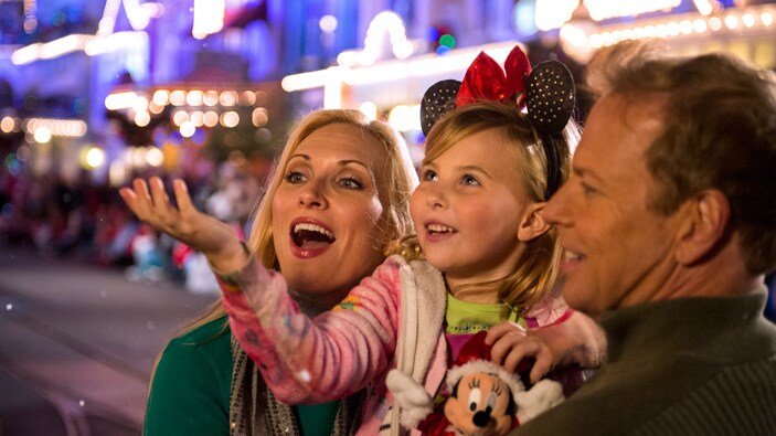 A girl dressed in Minnie Mouse ears, holding her hand out to catch snow while her parents watch