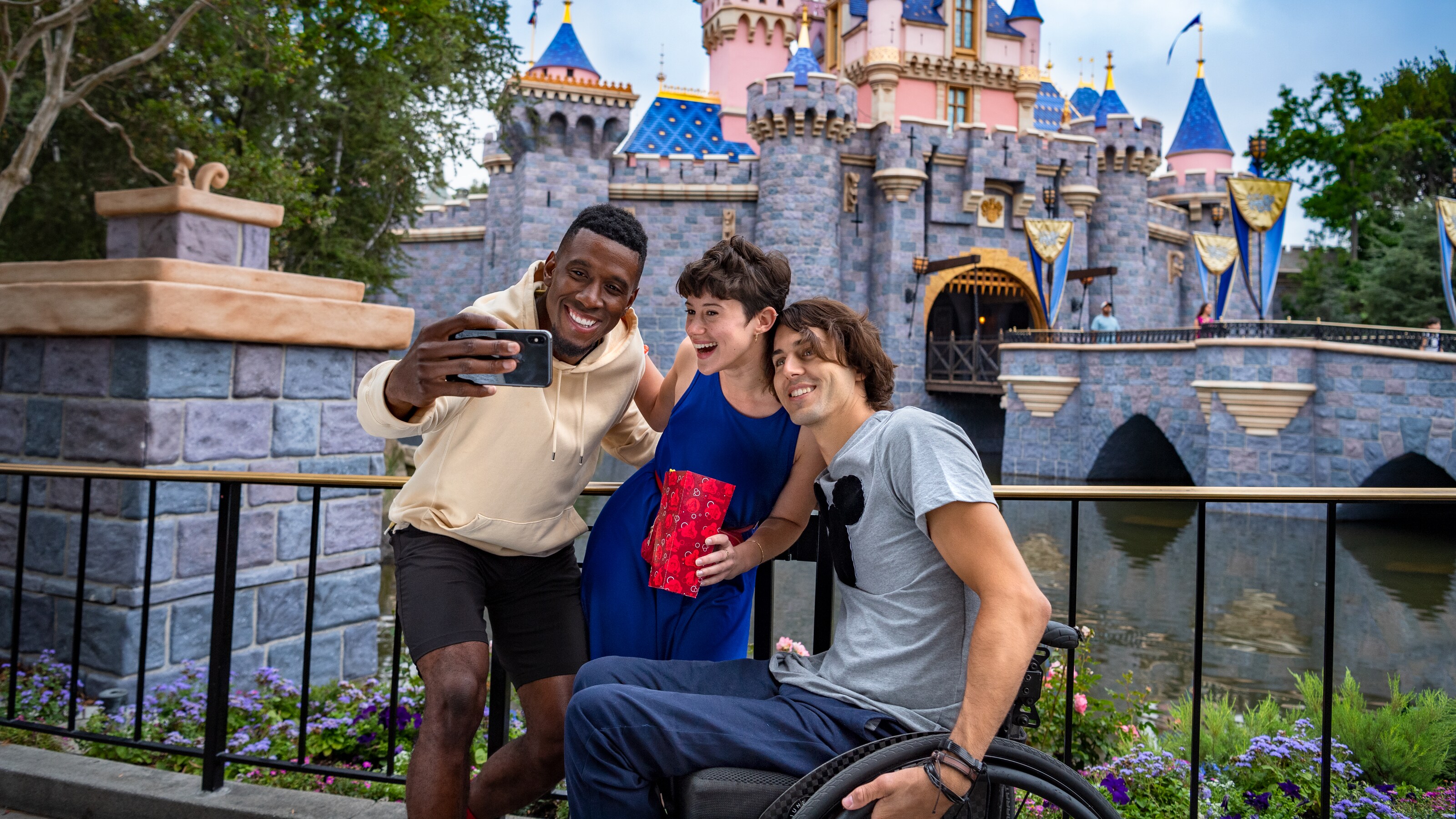 A man takes a picture with his phone of himself and 2 friends, 1 in a wheelchair, in front of Sleeping Beauty Castle in Disneyland park