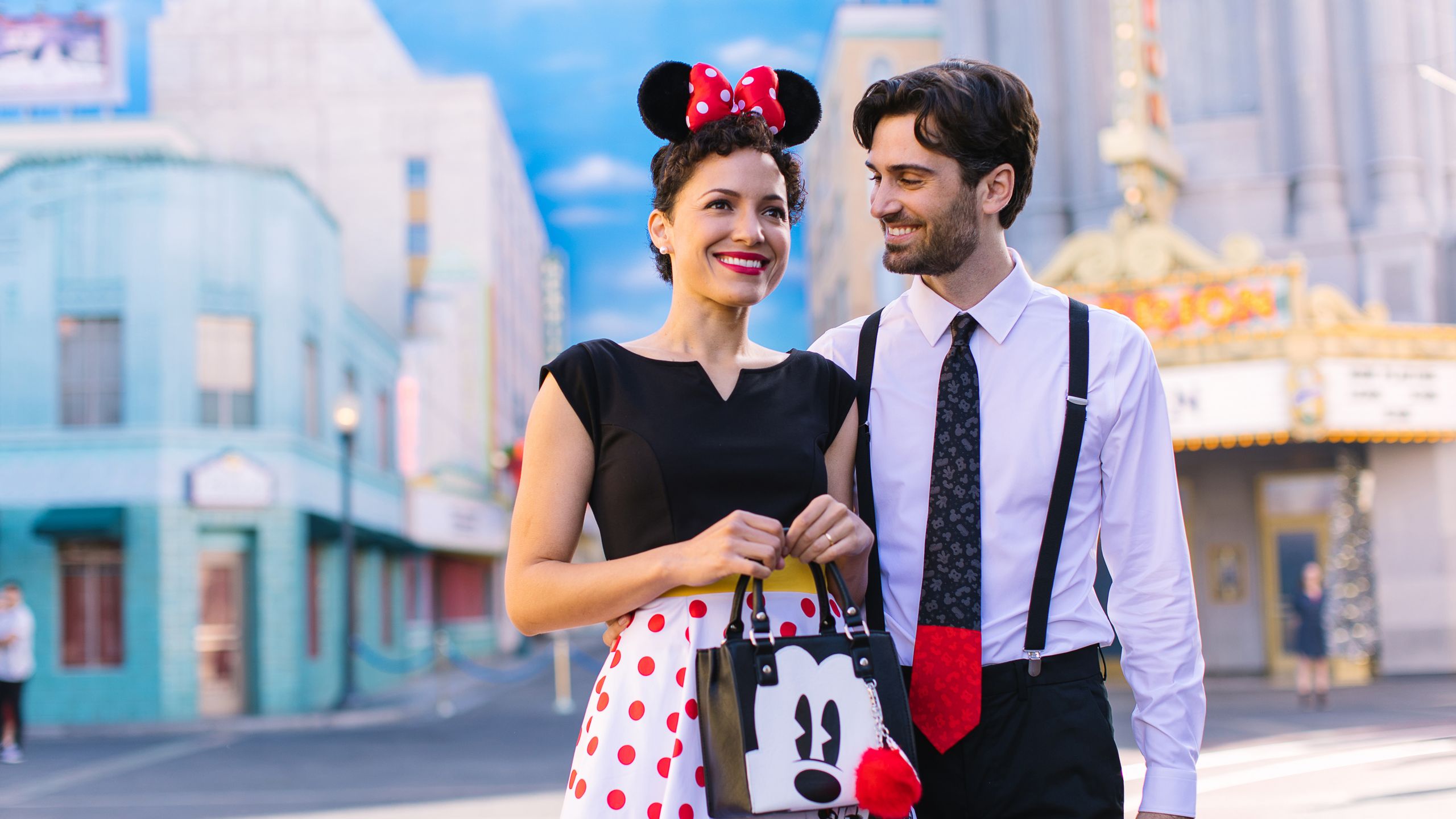 A woman wearing a polka dot dress and a Minnie Mouse ear headband walks through Hollywood Land with a man wearing a Mickey Mouse tie and suspenders in Disney California Adventure park at Disneyland Resort
