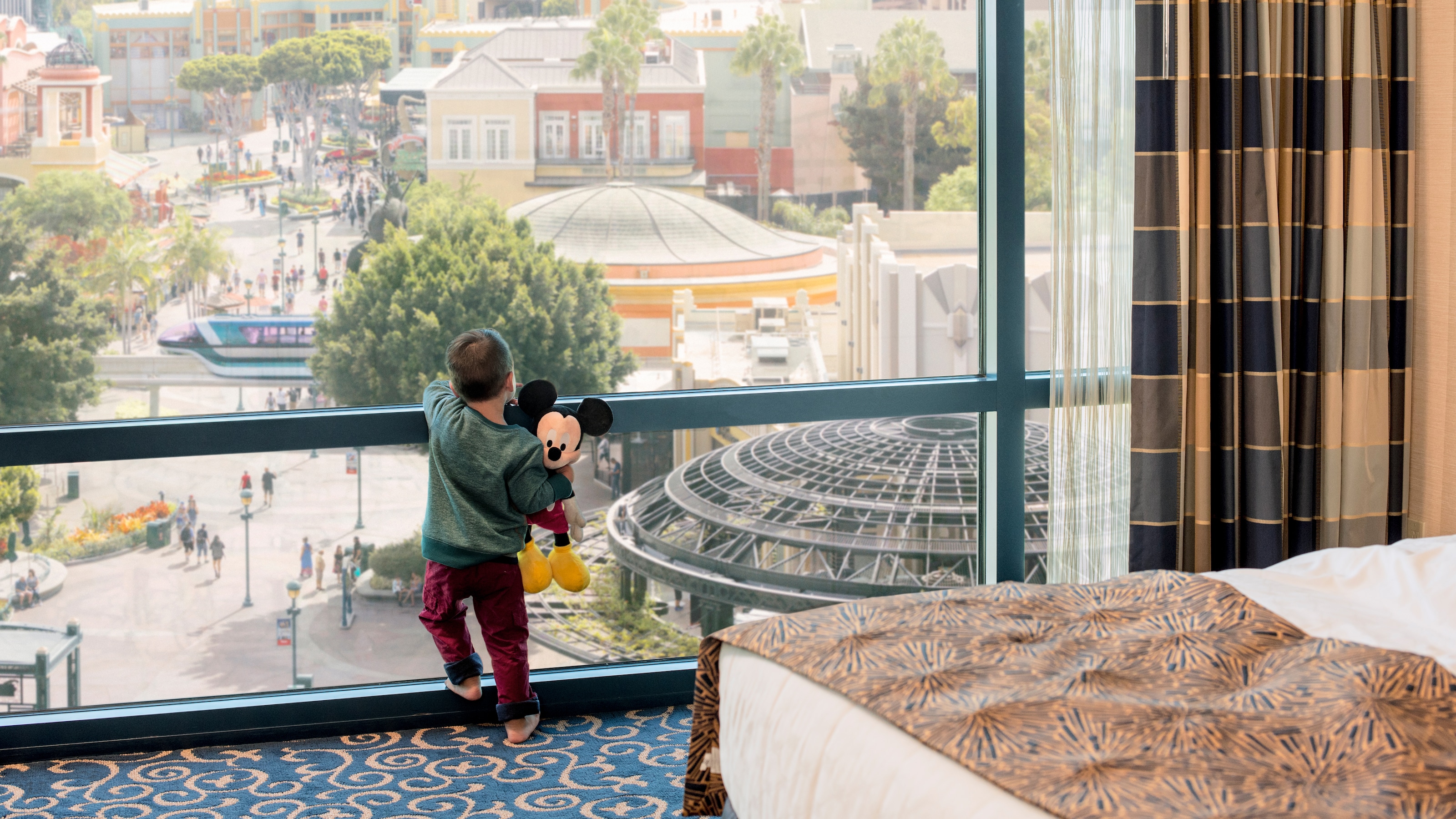 ミッキーマウスの人形を手に持ち、ホテル・ルームの窓からディズニーランド・パークを眺める小さな子ども