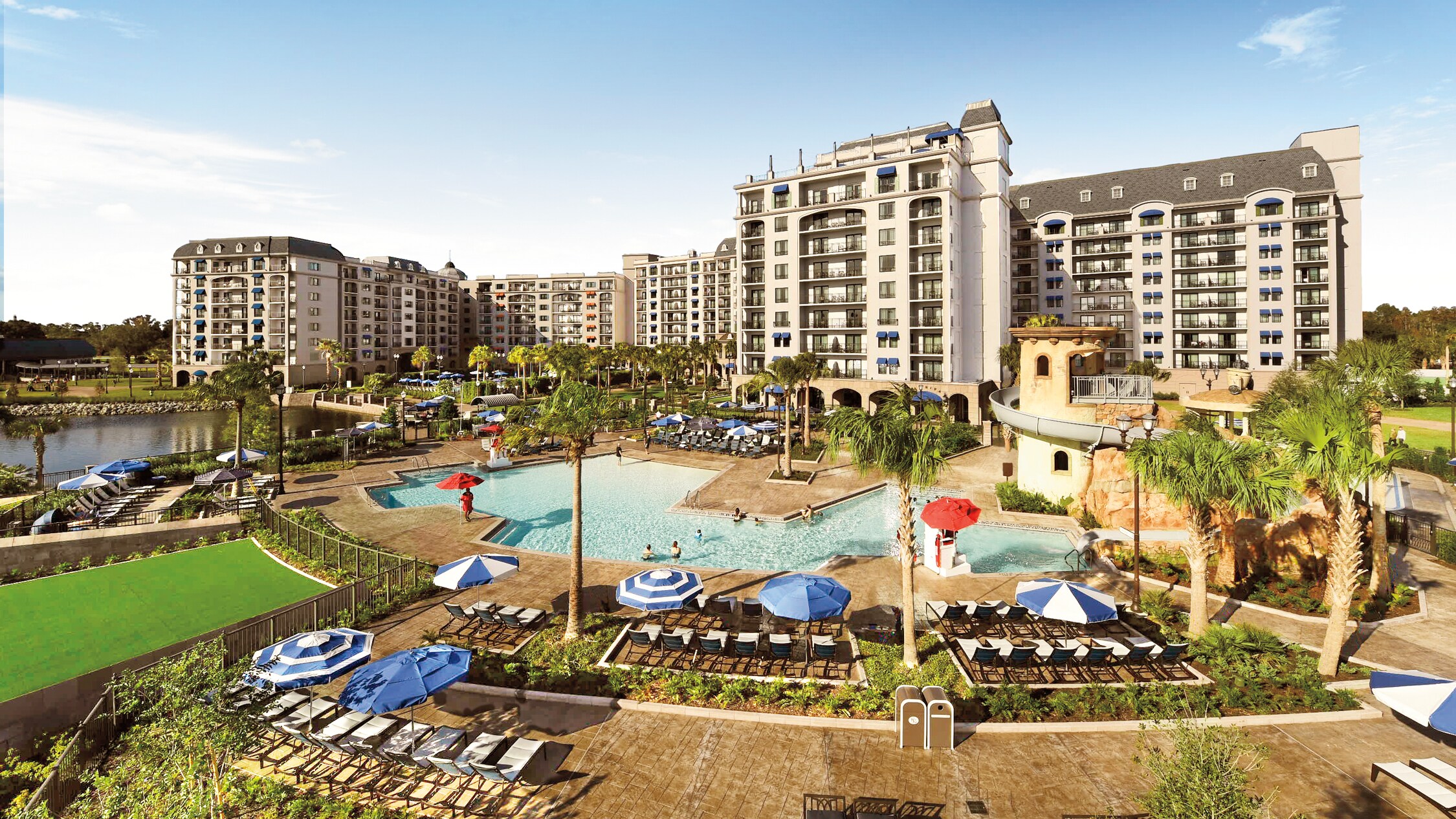 An outdoor pool with a waterslide, surrounded by lounge chairs, umbrellas and palm trees at Disney's Riviera Resort