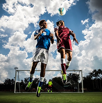 Two soccer players jumping to gain control of a soccer ball with blue sky and goal in the background.