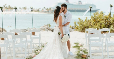 A man and a woman embracing and sharing a kiss in a tropical wedding setting with a Disney Cruise Line ship in the distance